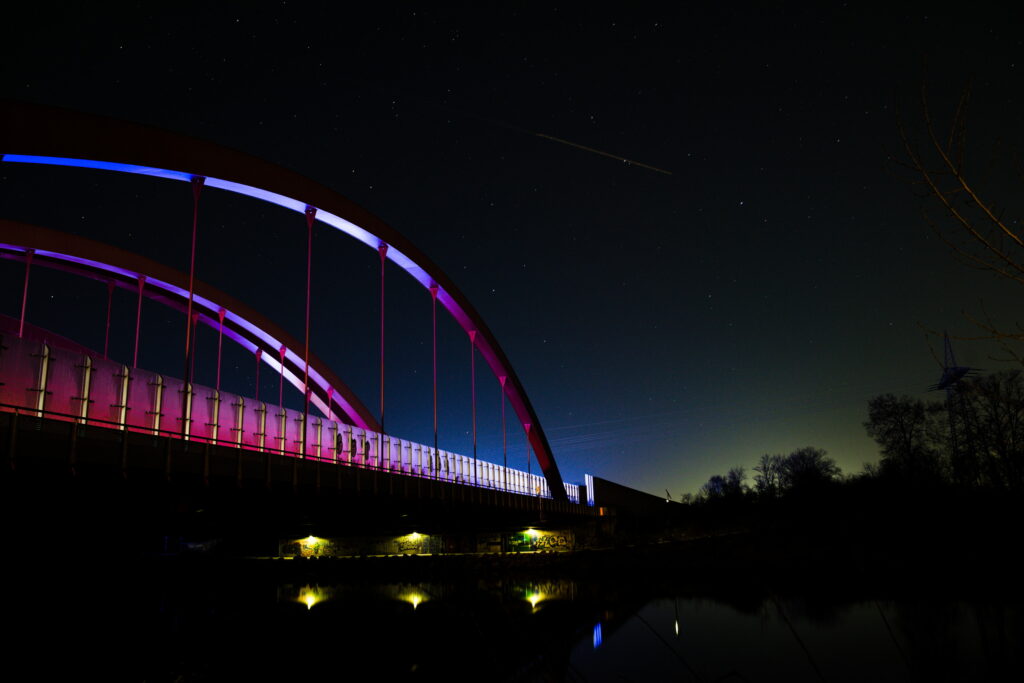 Eine Autobahnbrücke bei Nacht, beleuchtet durch vorbeifahrende Fahrzeuge, leuchtet in Hellblau, Rot und Blau. Bei Nacht.